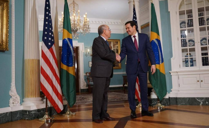 Dois homens de terno apertam as mãos em sala oficial decorada com bandeiras dos Estados Unidos e do Brasil. Lustres e móveis clássicos compõem o ambiente. Ministro das Relações Exteriores do Brasil, M